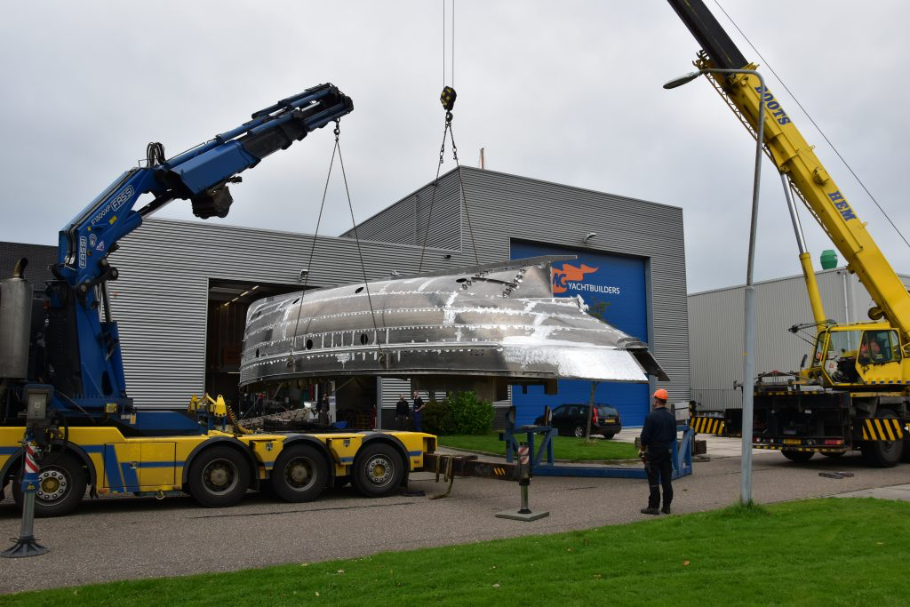 Twee kranen tillen het casco bij de werf van de transportwagen.