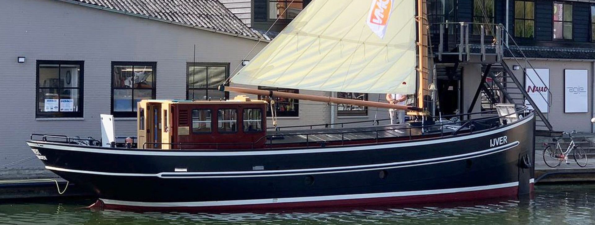 De IJver aan de kade, met de mast en het gehesen zeil boven de stuurhut.